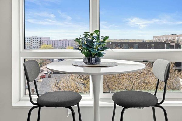 A room with a round table and two chairs, a potted plant on the table, and a view of buildings through a large window.
