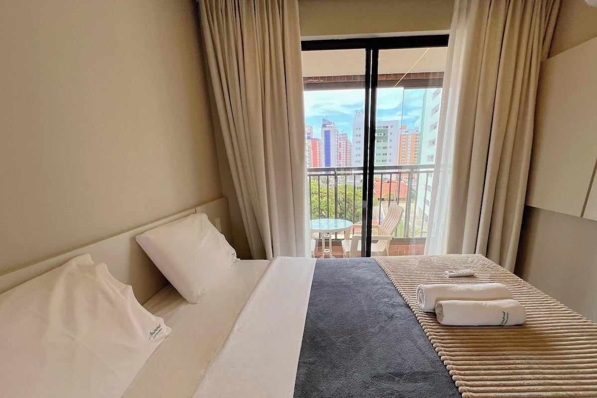 A hotel room with a bed, a view of the city through a balcony, and a window with curtains.