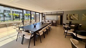 Uma sala de conferências moderna com uma mesa comprida, cadeiras brancas, uma tela de projetor e janelas amplas.