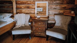 Une pièce rustique aménagée dans une cabane en bois, comprenant un lit, deux fauteuils, une petite table en bois avec un miroir et une commode.