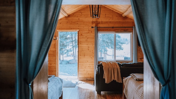 A cozy cabin interior with a wooden ceiling, a sofa with a blanket, and a view of a snowy landscape through a large window. - Aspen Cabin: Relaxing Mountain Escape | Sleeps 4 (Clearwater County)