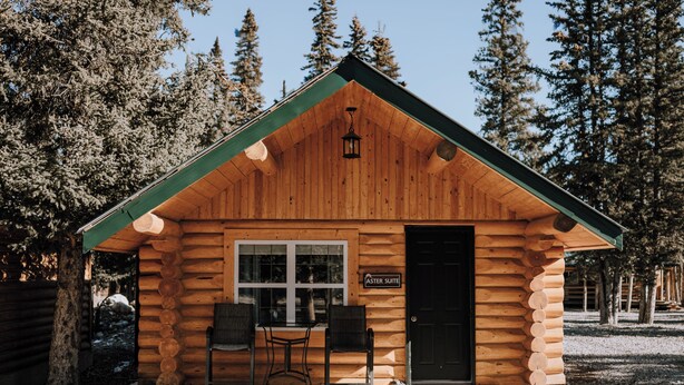 A log cabin with a green roof, a wooden door, and two chairs outside.
