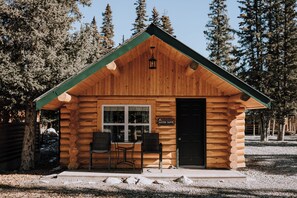 Une cabane en rondins avec un toit végétalisé, une porte portant l’inscription « Suite Aster », et deux chaises à l’extérieur.
