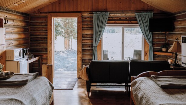 Habitación de cabaña con dos camas, techo de madera, una pequeña cocina y una ventana con cortinas.