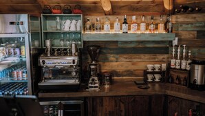 Una zona de bar con un refrigerador lleno de bebidas, una cafetera y estantes con botellas y vasos.