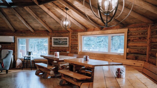 Une cabane en bois rustique, dotée d’une grande table à manger, de bancs et d’un lustre.