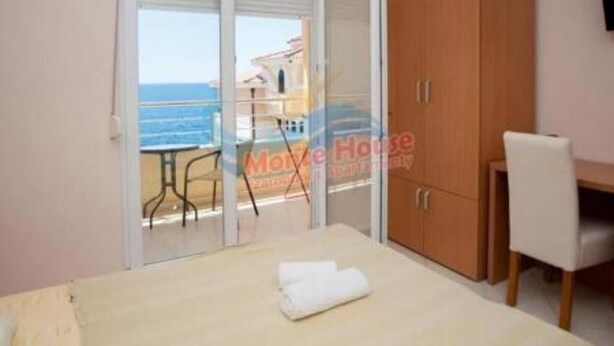 Une chambre à coucher comprenant un lit, une armoire en bois, un bureau et une chaise. Une porte-fenêtre coulissante donne accès à un balcon avec vue sur la mer.