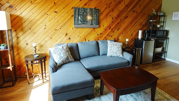 A living room with a wooden wall, a gray sofa, a wooden coffee table, and a kitchen area with a microwave. - Homestead Motel & RV (Sloan)