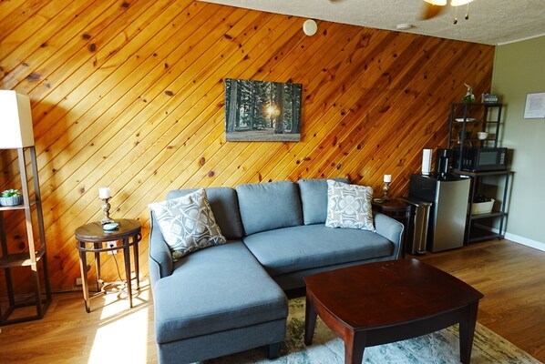 A living room with a wooden wall, a gray sofa, a wooden coffee table, and a kitchen area with a microwave.