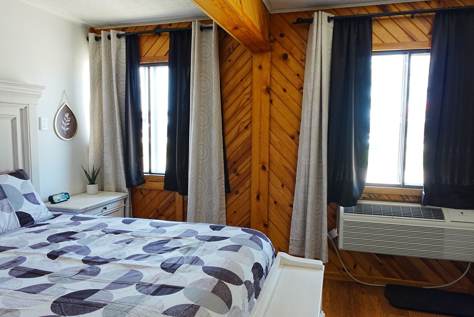 A bedroom with a bed, a nightstand, a potted plant, a wall-mounted heater, and wooden paneling.