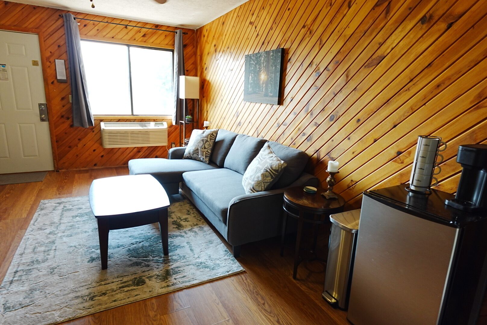 A room with a sofa, a small table, a lamp, a coffee maker, and a wall-mounted TV.