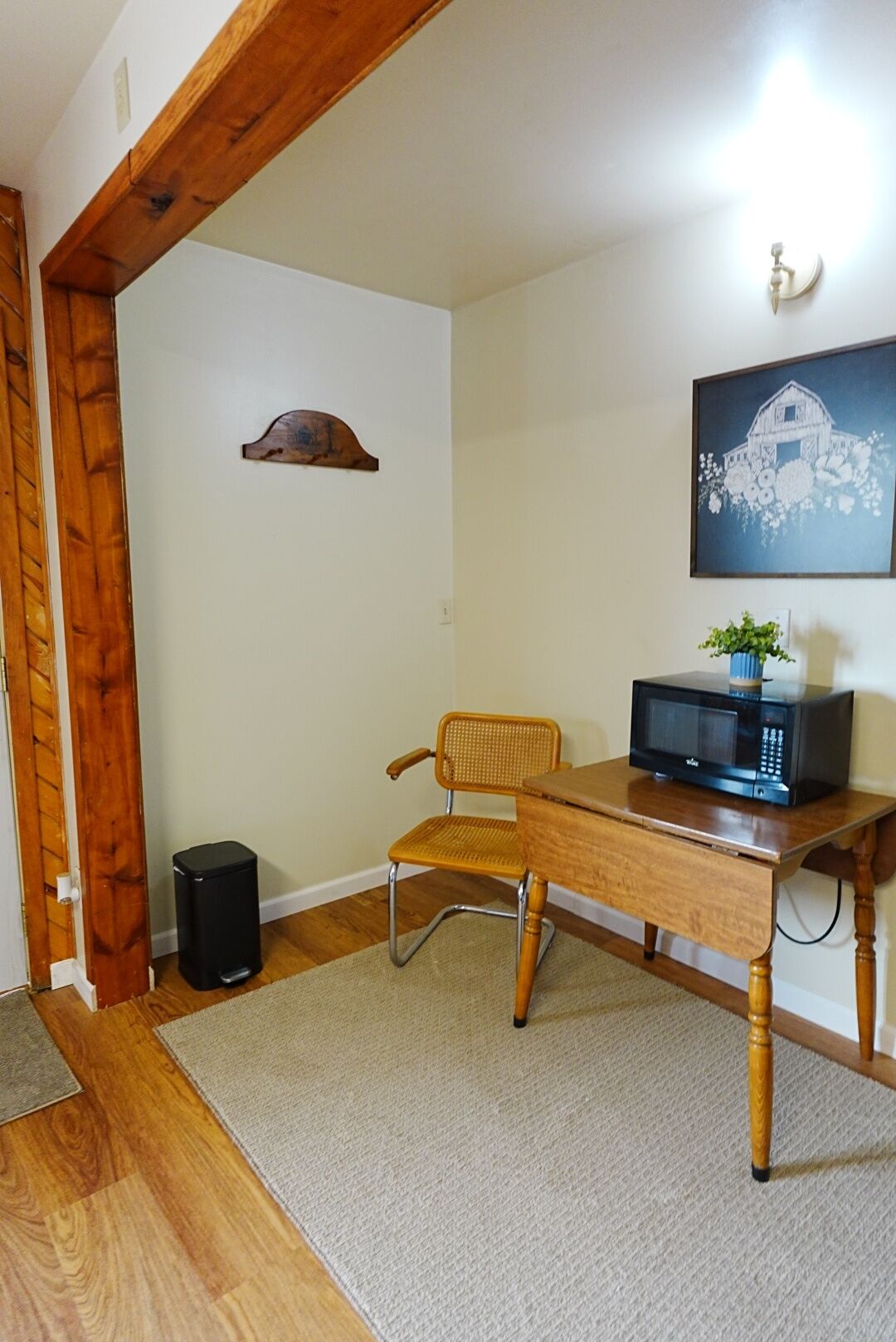 A room with a wooden desk, a chair, a trash bin, a microwave, and a framed picture on the wall.