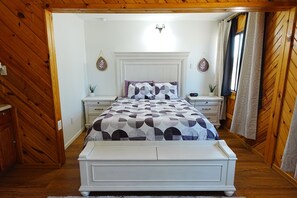 A bedroom with a bed, bedside tables, a nightstand, a wall-mounted light fixture, and a window with curtains. - Homestead Motel & RV (Sloan)
