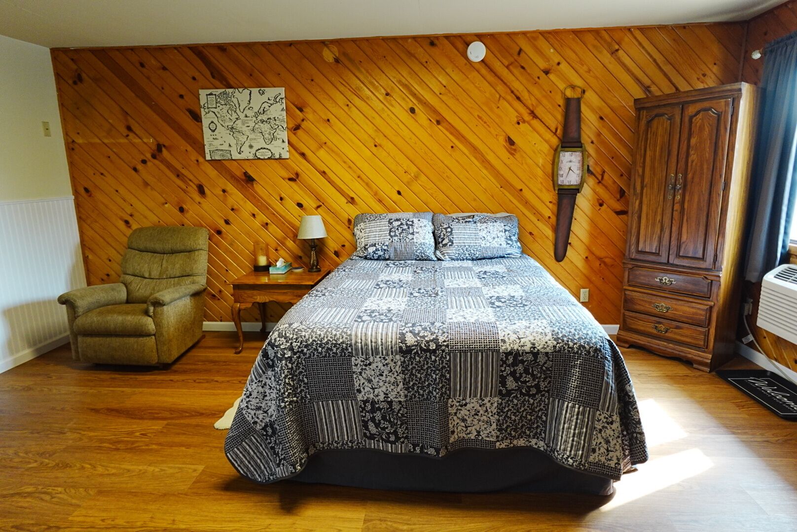 A bedroom with a wooden wall, a bed with patterned bedding, a wooden chair, a nightstand with a lamp, and a wardrobe.