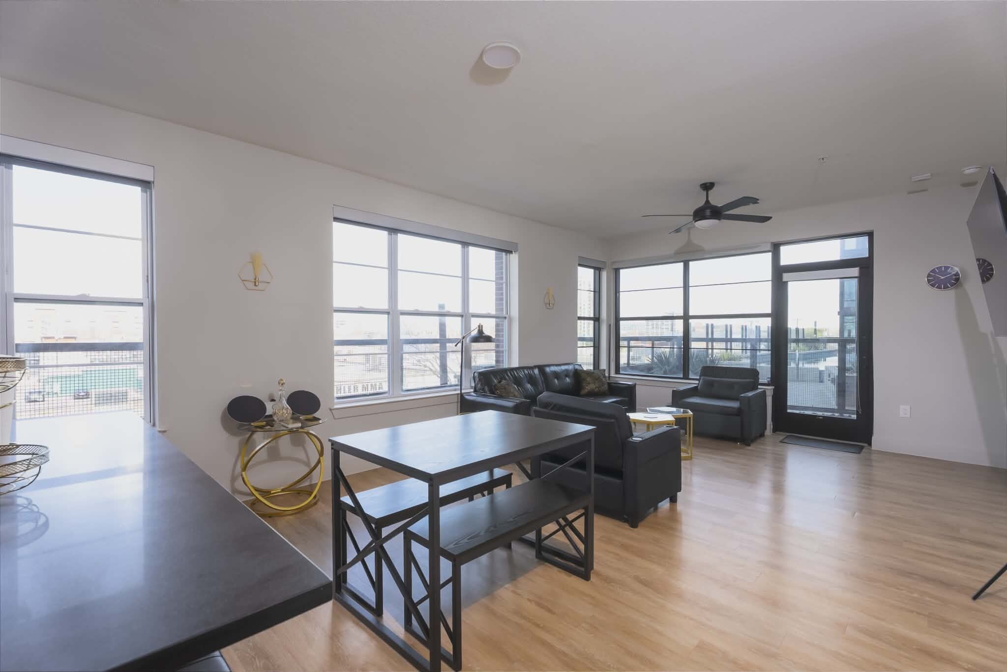 A spacious living area with a dining table, sofa, and chairs. Large windows provide ample natural light and views of the city.