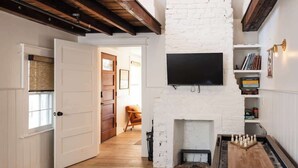 A room with a white brick wall, a fireplace, a flat-screen TV mounted above it, and a wooden floor. - The Grand Helène - Newly Renovated 11BD, 12BA - 2 Blocks from King St! (Charleston)