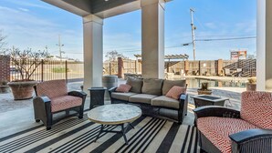 A covered outdoor seating area with wicker furniture, a round table, and a view of a parking lot and buildings in the background.