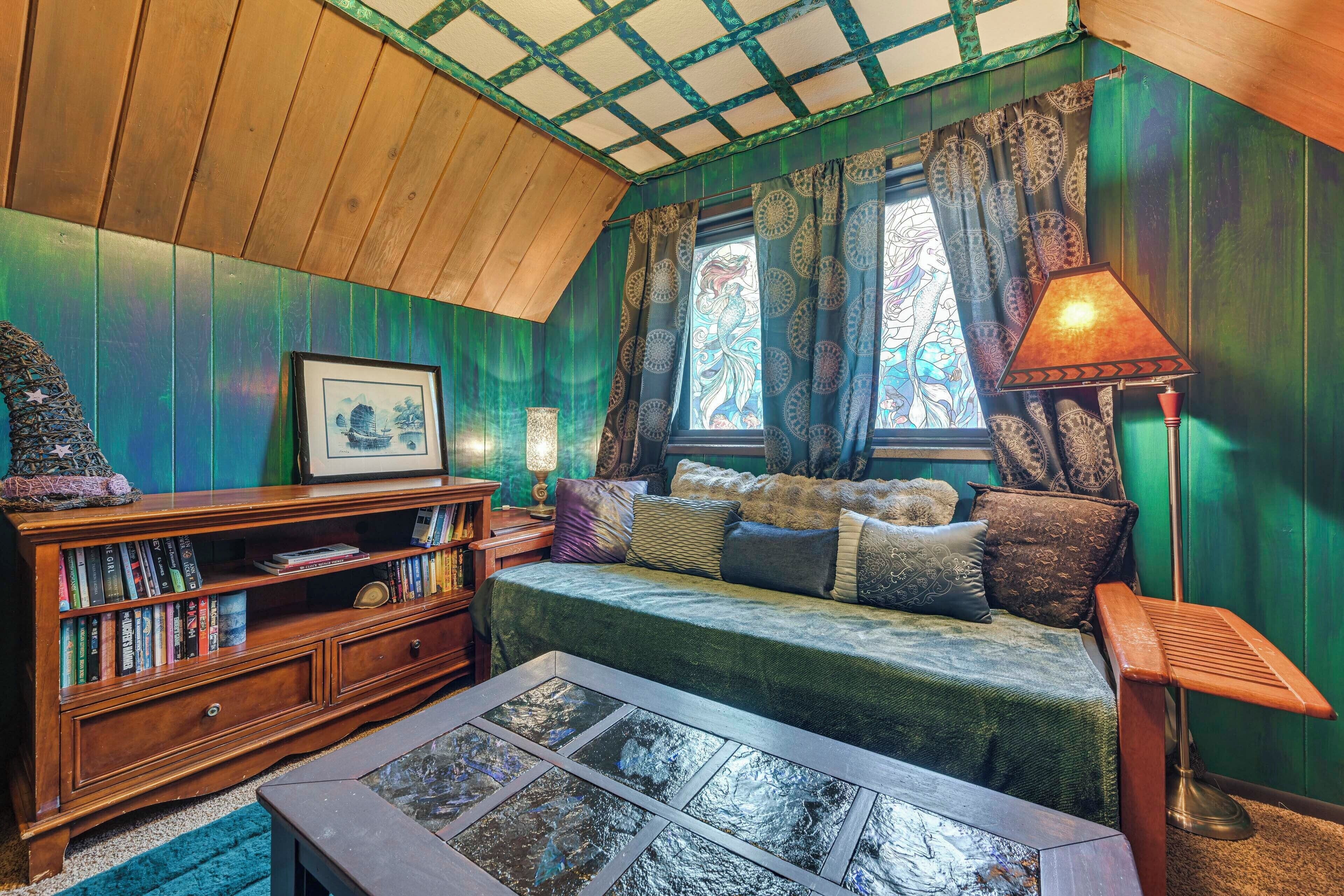A cozy room with a wooden ceiling, a green wall, a sofa with cushions, a bookshelf, and a window with patterned curtains.