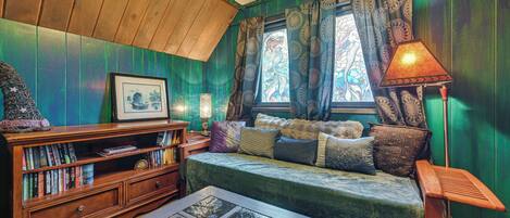 A cozy room with a wooden ceiling, a green wall, a sofa with cushions, a bookshelf, and a window with patterned curtains.
