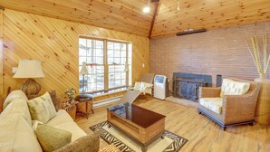 A cozy living room with a wooden ceiling, a brick fireplace, a sofa, a wicker armchair, and a glass coffee table. - 1 Mi to TIEC: Peaceful Mountain Cabin (Mill Spring)