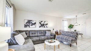 Ein modernes Wohnzimmer mit einem grauen Ecksofa, einem gemusterten Sessel und einem weißen Couchtisch. An der Wand befinden sich abstrakte Gemälde und im Hintergrund ein Essbereich.