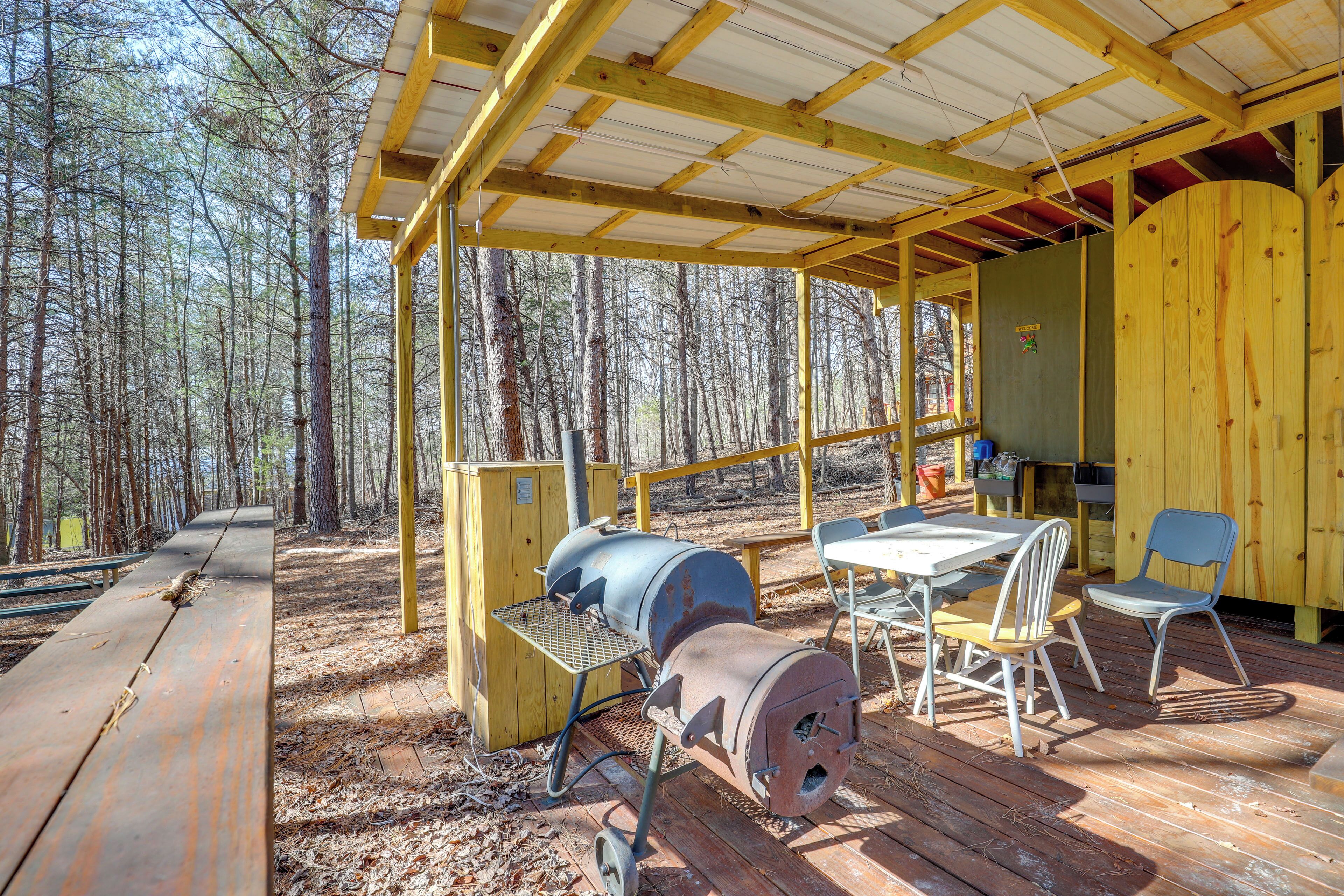 Ein überdachter Außenbereich mit Holzterrasse, Grill, Tisch und Stühlen sowie Blick auf den umliegenden Wald.