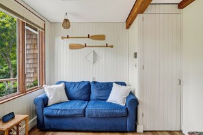 A room with a blue sofa, wooden beams, a window with blinds, a wall-mounted paddleboard, and a small table with a clock. - Steps to Beach: Martha's Vineyard Private Retreat! (Vineyard Haven)