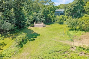 Ein grasbewachsenes Gelände mit einer Holzterrasse und einem Picknicktisch, umgeben von dichten Bäumen und einem Haus im Hintergrund.