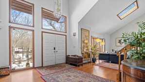 Un salón espacioso con pisos de madera, ventanas amplias, un sofá oscuro, una mesa de centro de madera y un tapete grande.