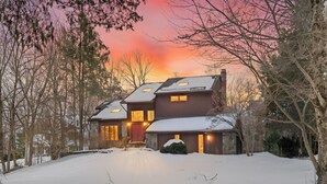 Una casa de dos pisos con un camino de entrada cubierto de nieve y un atardecer en el fondo.