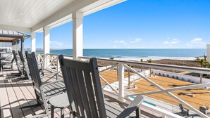A covered deck with wooden chairs and tables, overlooking a beach and ocean. - Paradise at the Pointe (Ocean Isle Beach)