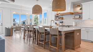 A modern kitchen with a large island, wooden chairs, pendant lights, and white cabinetry. - Paradise at the Pointe (Ocean Isle Beach)