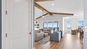 A spacious living area with a high ceiling and wooden beams, a seating area with sofas and armchairs, and a view of the sea through large windows. - Paradise at the Pointe (Ocean Isle Beach)