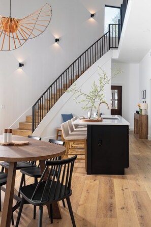 A modern kitchen with a central island, a dining area with a wooden table and chairs, a staircase with black railings, and a view of the outdoors. - Luxe Silo Retreat: Pickleball, Sauna, Hot Tub (Fennville)