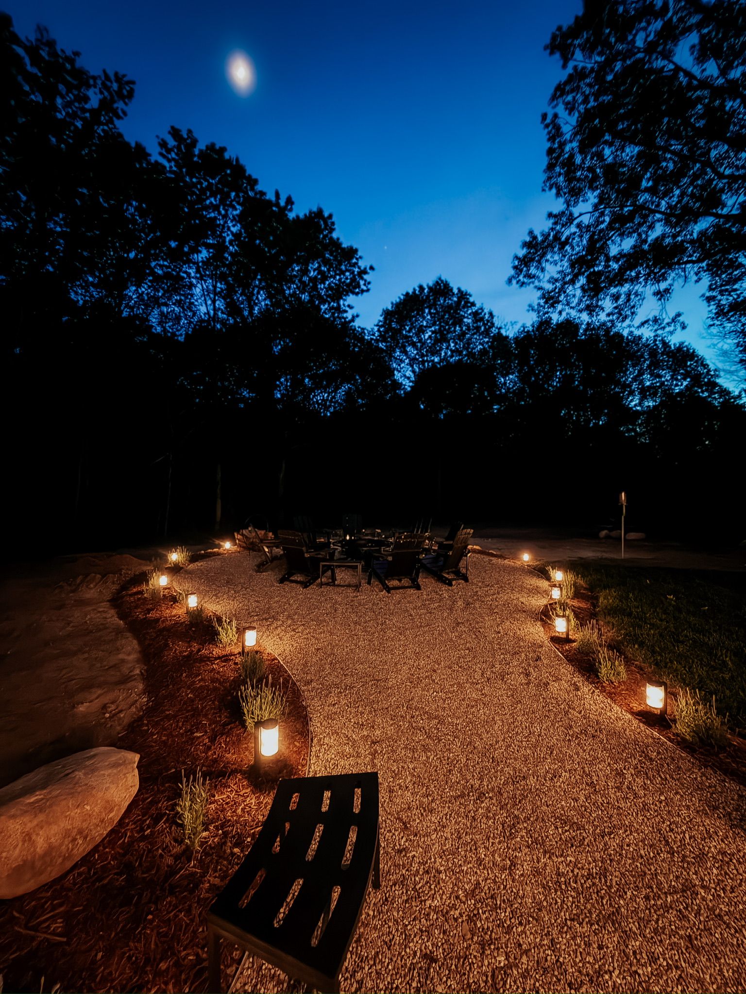 Un espace extérieur aménagé avec des sièges, un chemin de gravier, entouré d’arbres et illuminé par de petites lumières.