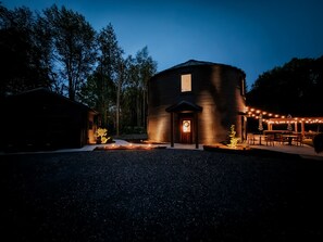 A rustic building with a circular design, a front door with a wreath, and a patio area with string lights. - Luxe Silo Retreat: Pickleball, Sauna, Hot Tub (Fennville)