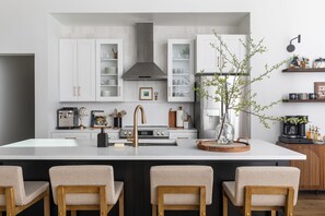 A modern kitchen with white cabinetry, a central island with bar stools, a stainless steel range hood, and a coffee machine. - Luxe Silo Retreat: Pickleball, Sauna, Hot Tub (Fennville)