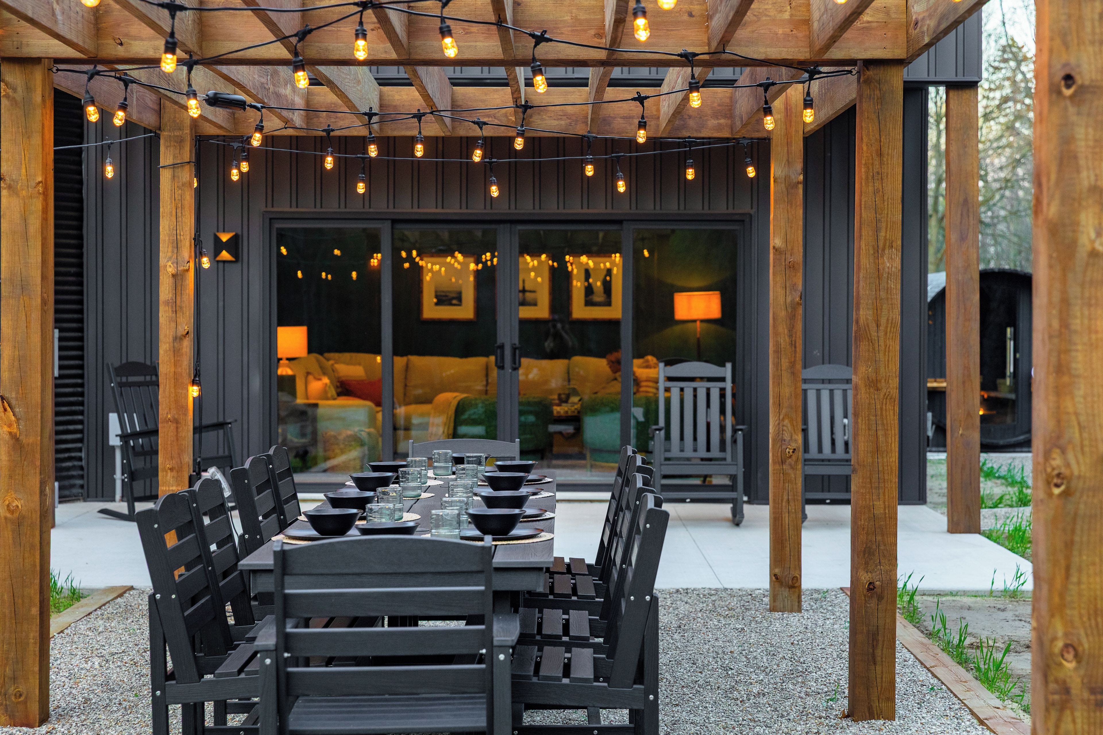 Een buitenrestaurant met een houten pergola, lichtslingers en uitzicht op een gebouw met een donkere buitenkant en binnenkant.