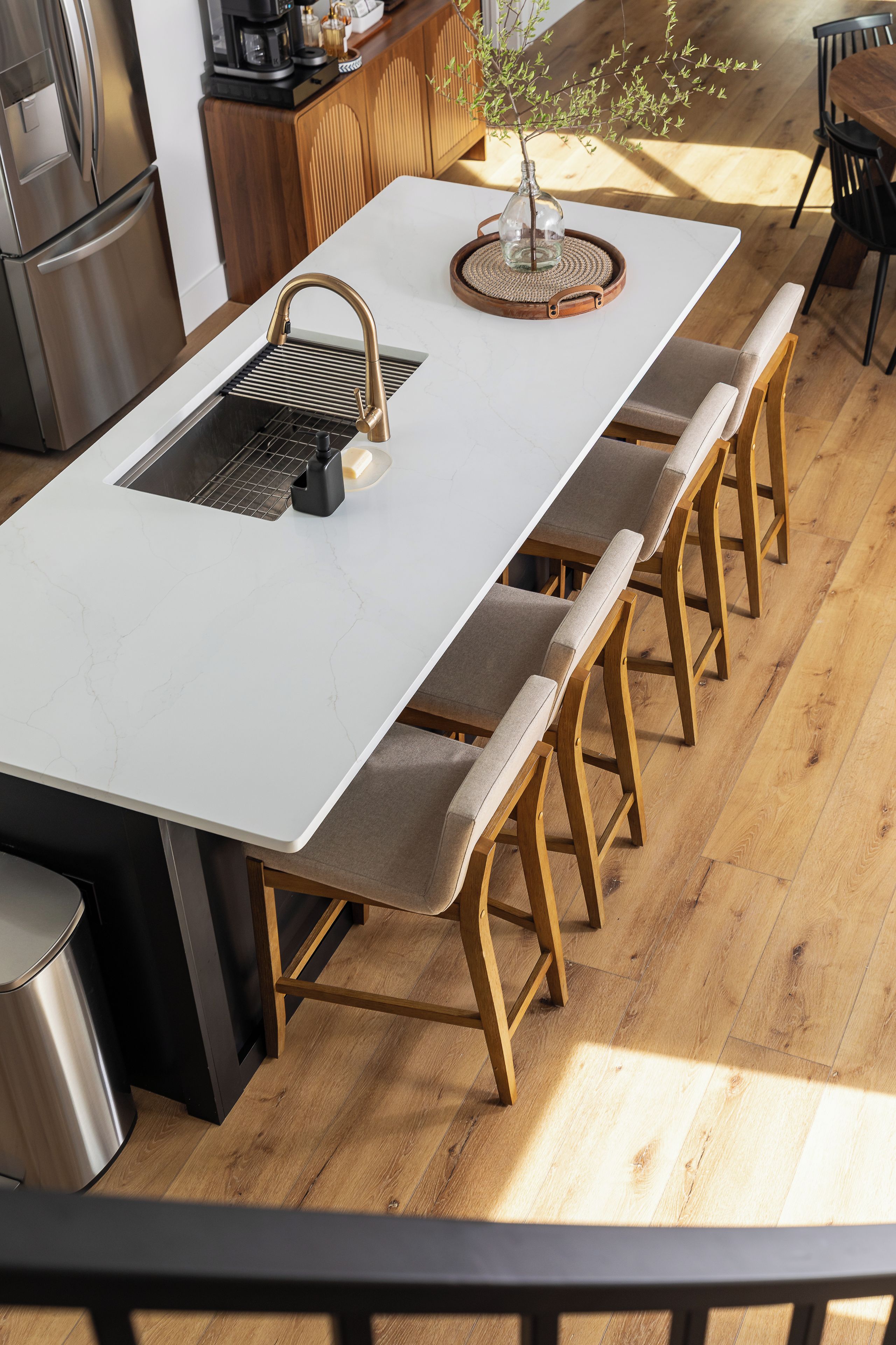 Une cuisine moderne avec un plan de travail blanc, des appareils en acier inoxydable, des tabourets en bois et une petite plante en pot.