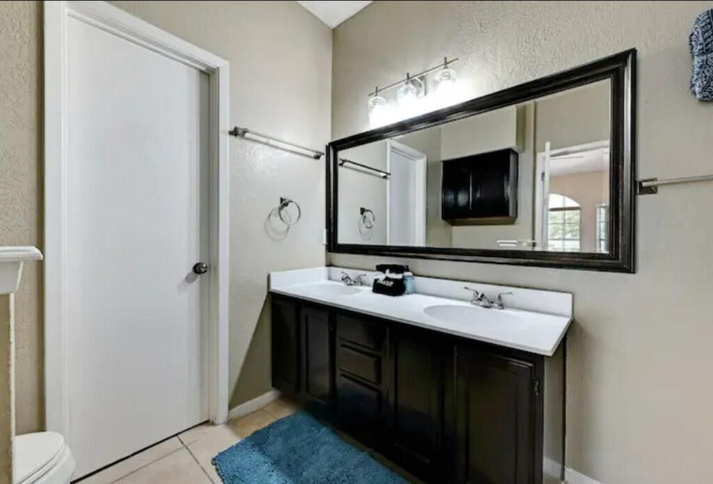 A bathroom with a white toilet, a double sink vanity, a large mirror, and a blue rug on the floor.