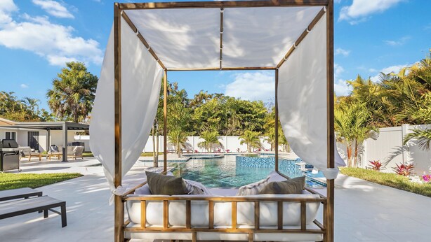 Una cama con dosel de madera con cojines blancos, con vistas a una piscina y abundante vegetación.