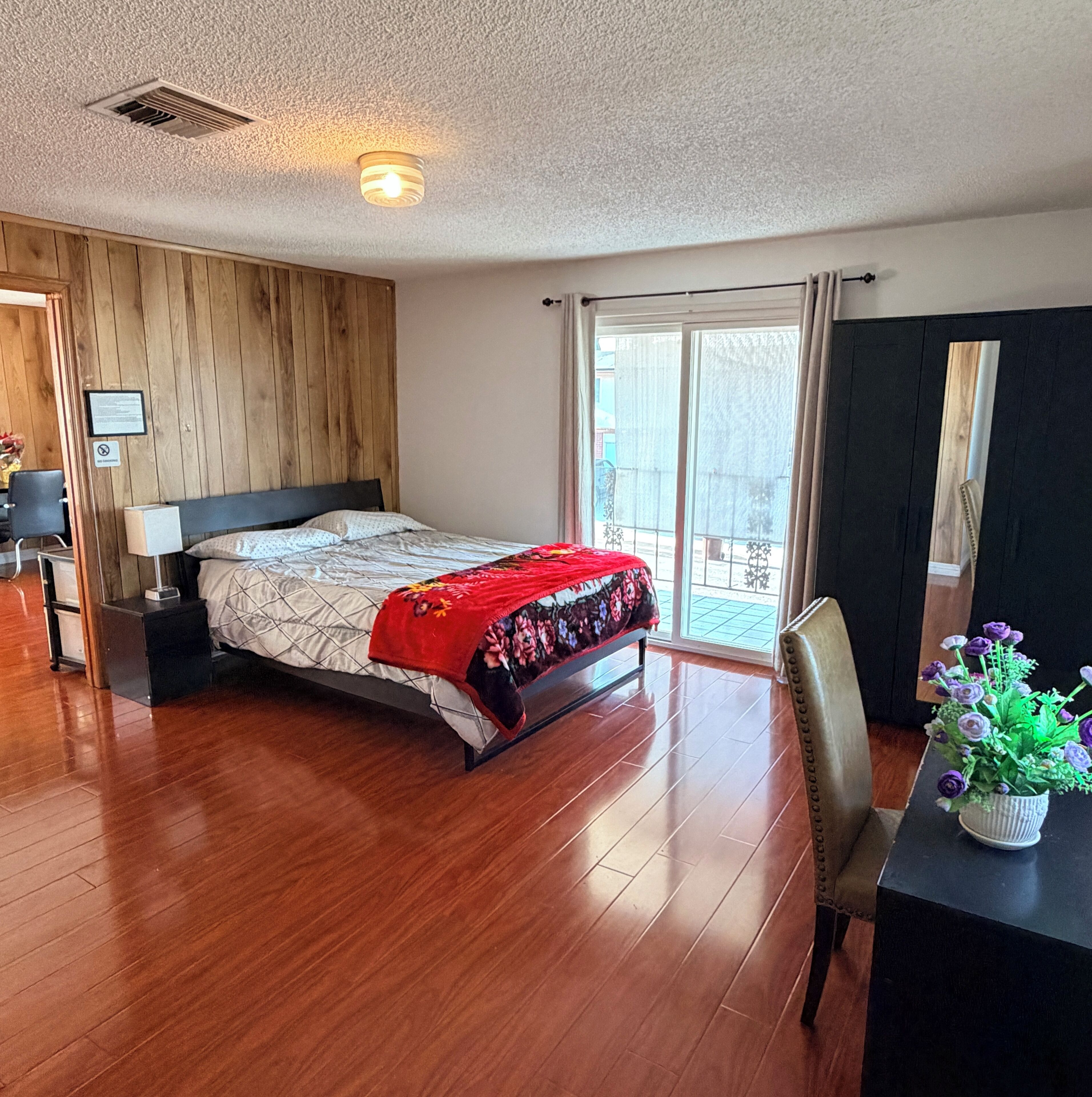 Quarto com cabeceira de madeira, cama com manta vermelha, mesa de cabeceira, escrivaninha com planta em vaso e porta de vidro deslizante para o exterior.