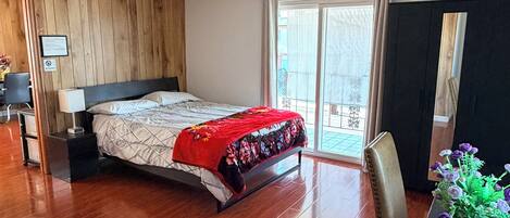 Quarto com cabeceira de madeira, cama com manta vermelha, mesa de cabeceira, escrivaninha com planta em vaso e porta de vidro deslizante para o exterior.