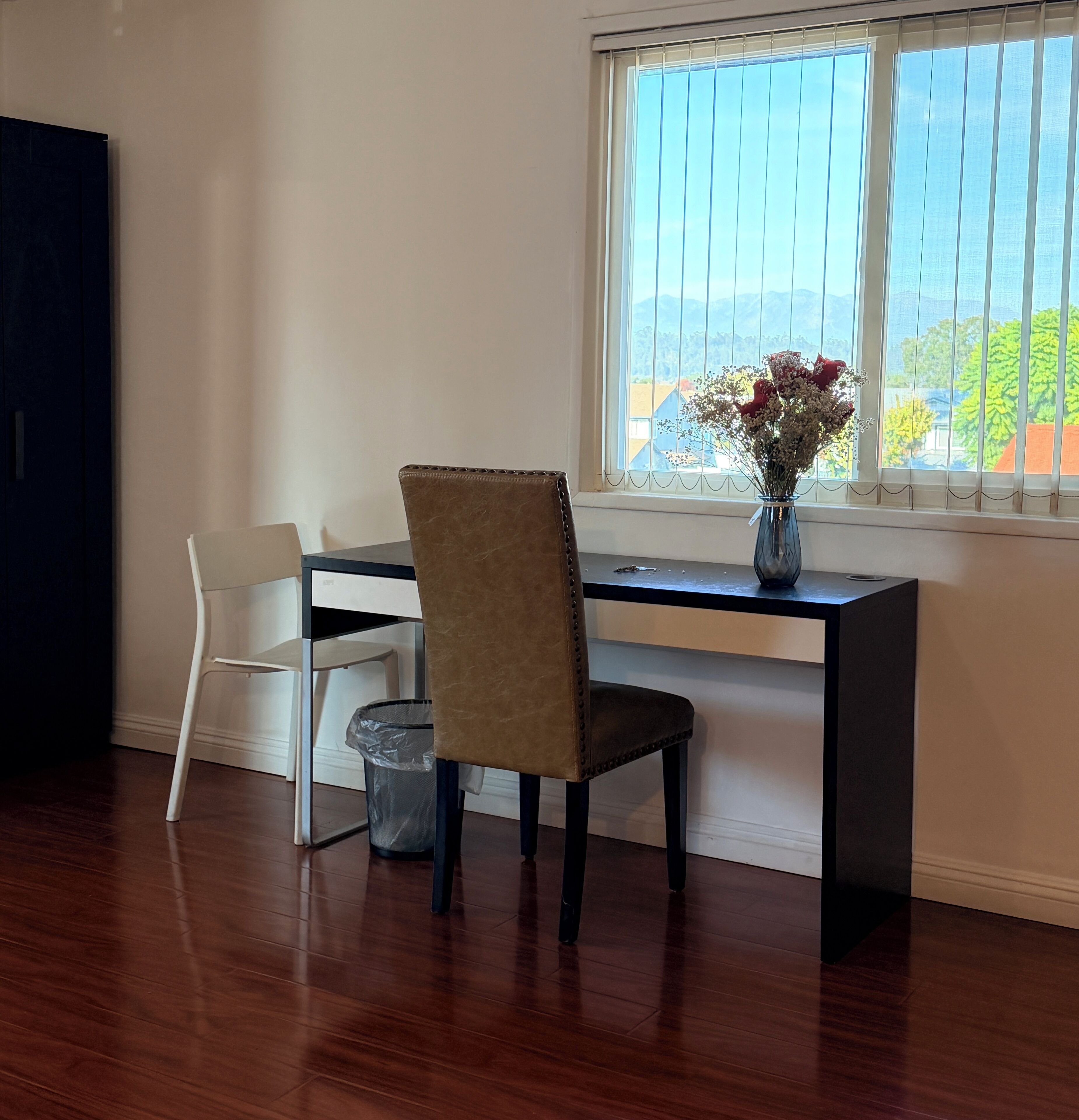 Ein Zimmer mit Holzboden, einem Schreibtisch mit Stuhl, einer Vase mit Blumen und einem Fenster mit Jalousien.
