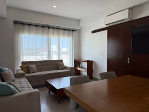 Una sala moderna con sofá, mesa de centro y mesa de comedor. Hay una ventana con cortinas translúcidas, un estante empotrado y un equipo de aire acondicionado en la pared.