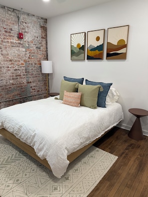 A bedroom with a brick wall, a bed with white bedding, and three framed artworks above the bed. - A Modern Coastal Escape in the Heart of Downtown (Wilmington)