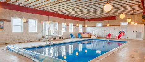 An indoor swimming pool with a clear blue water, surrounded by red and blue chairs, and a wooden ceiling with hanging lights.