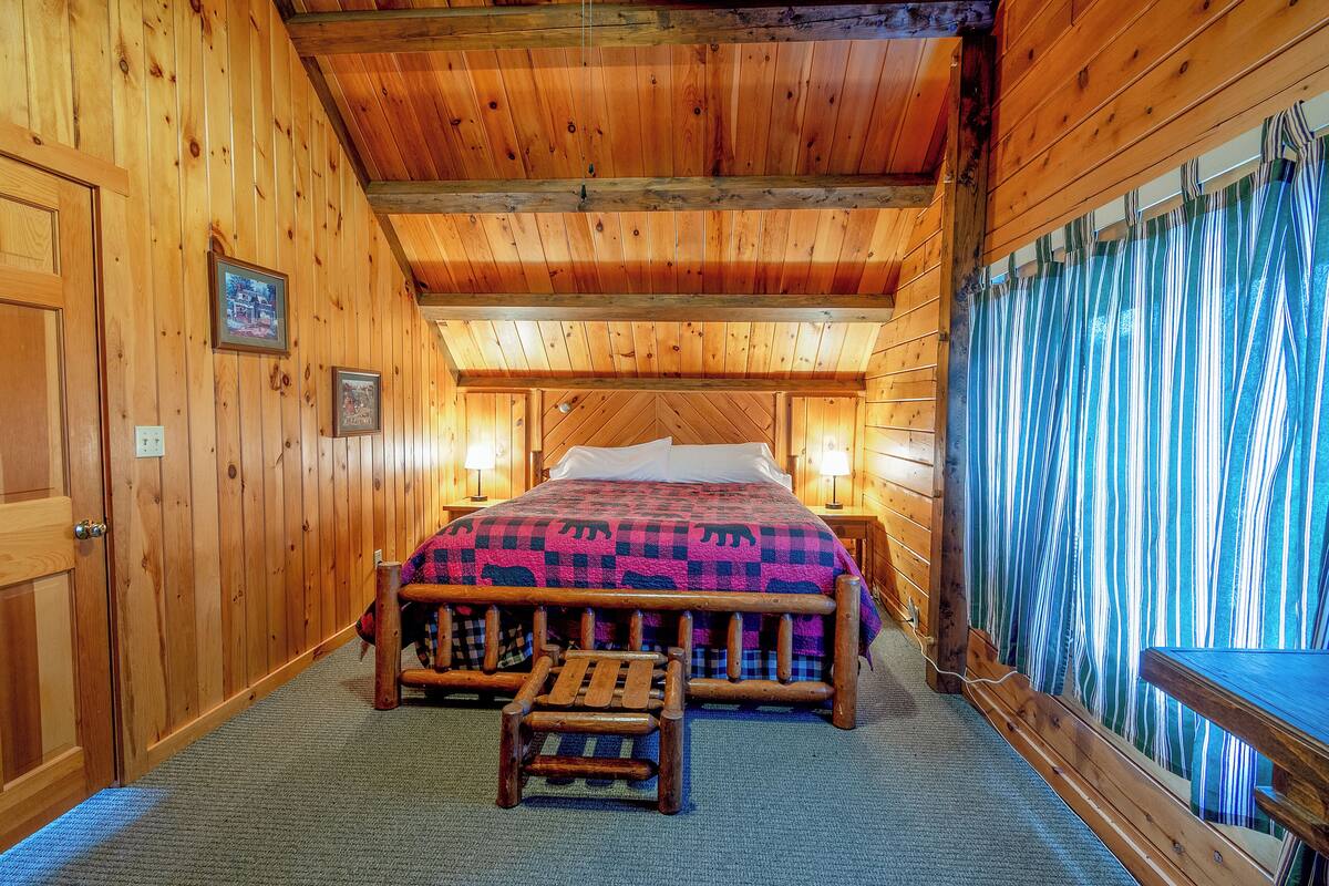 A wooden cabin bedroom with a bed, a bench, and a view of a forest through a window.