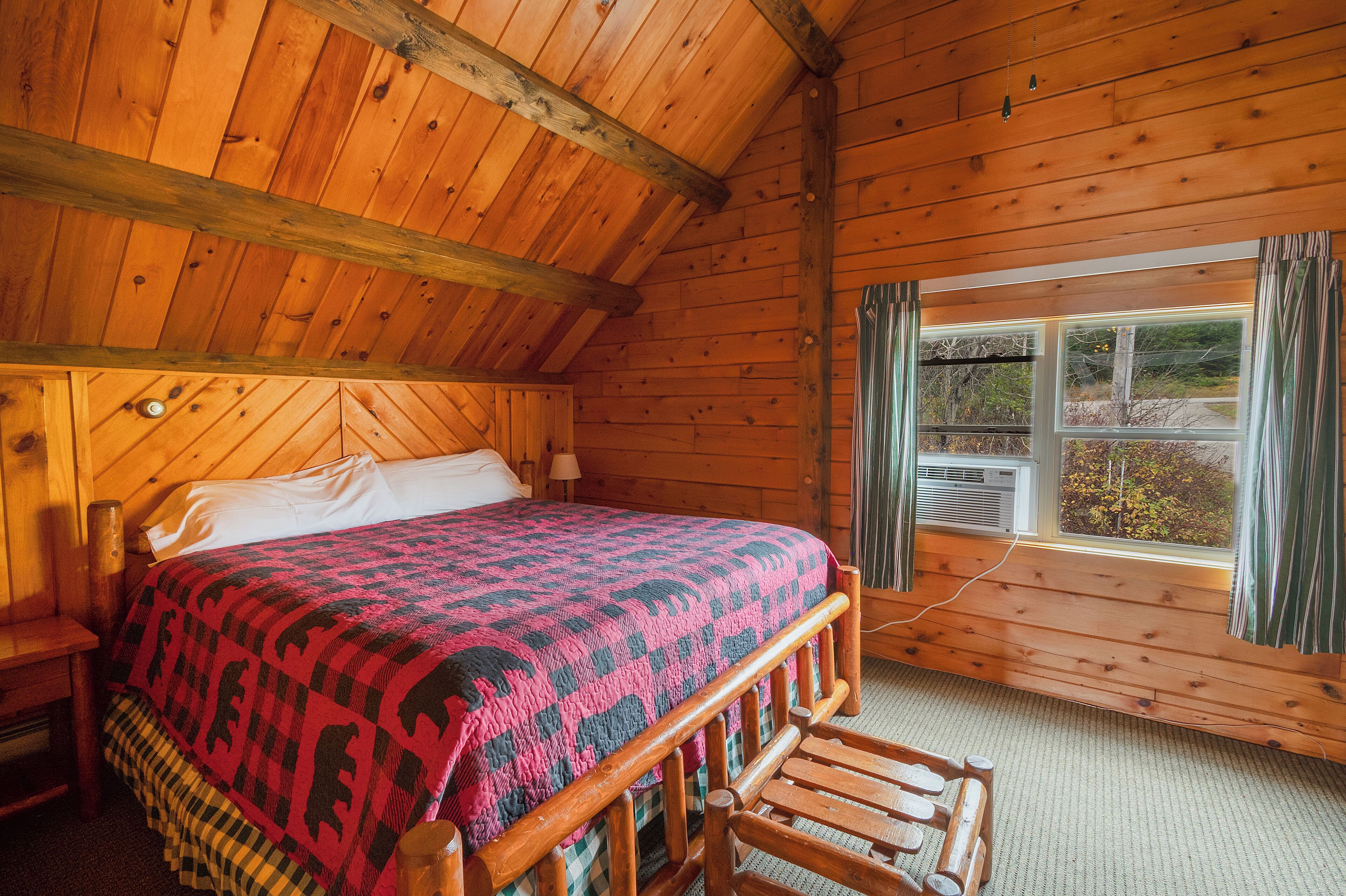 A wooden cabin bedroom with a bed, a window with curtains, and a wooden headboard.