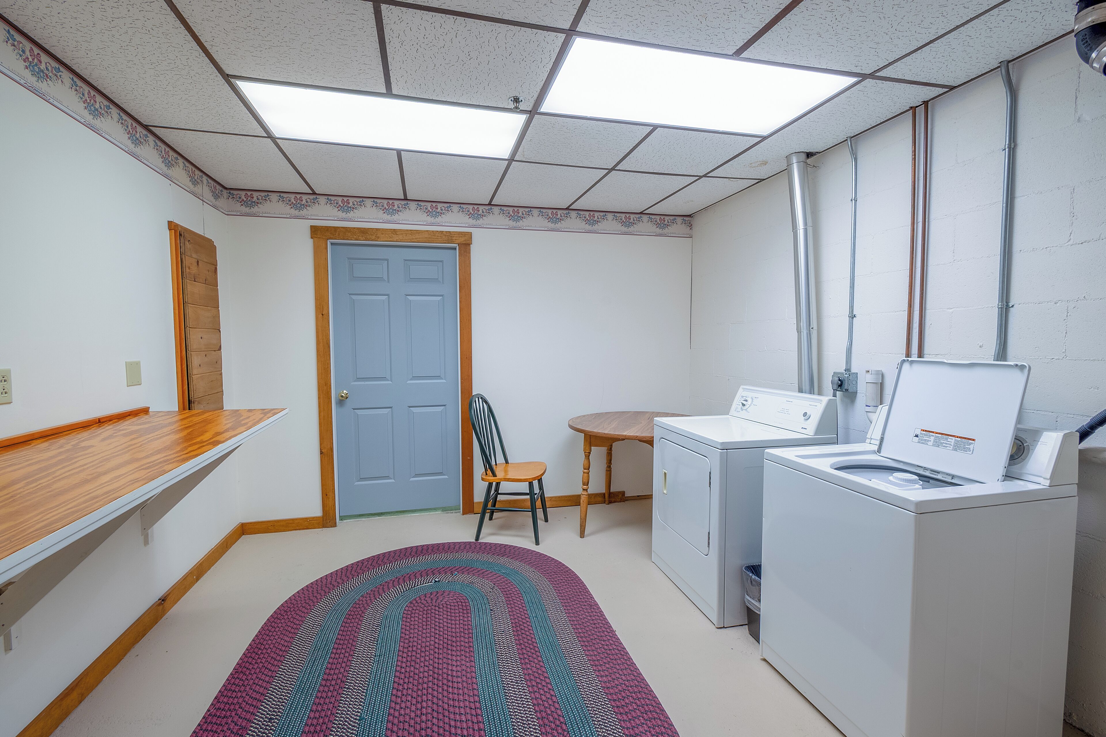 A laundry room with a washing machine, a dryer, a chair, a small table, and a door.